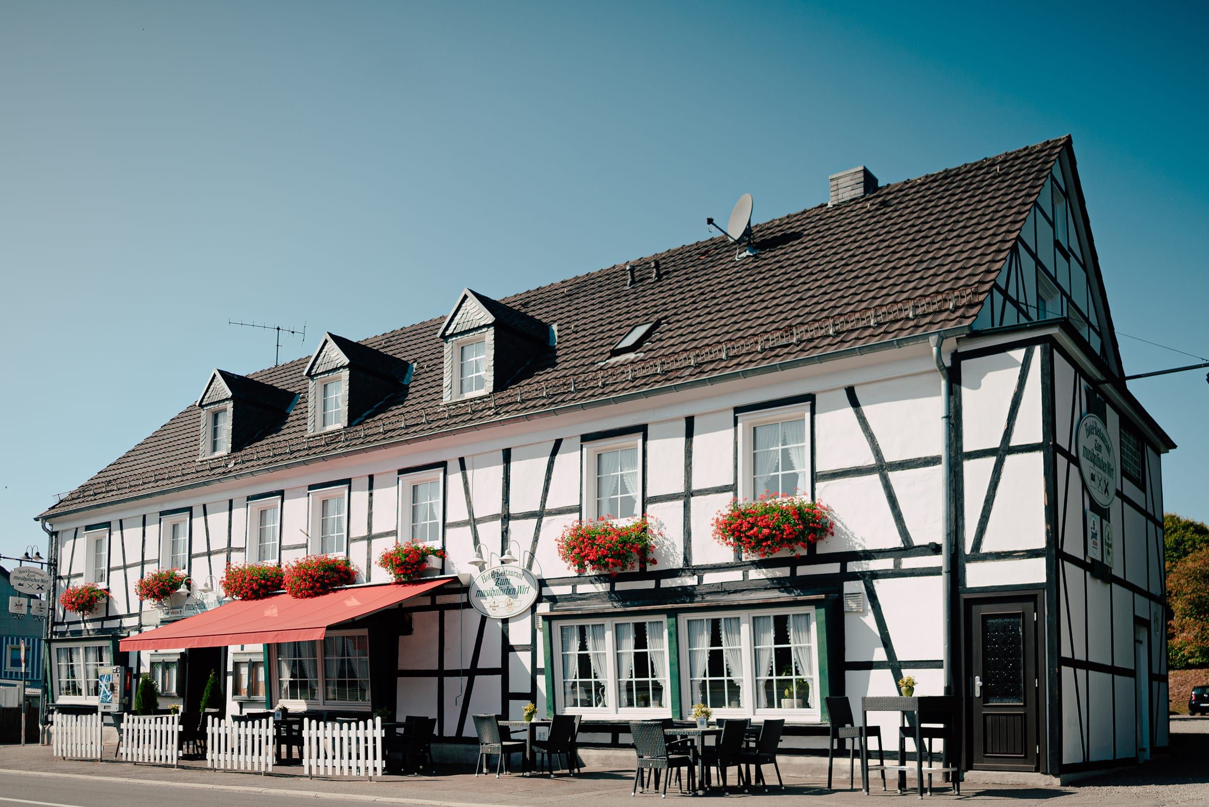 Sonnenbeschienene Fachwerkfassade des Hauses mit roter Markise, Geranien und Terrasse